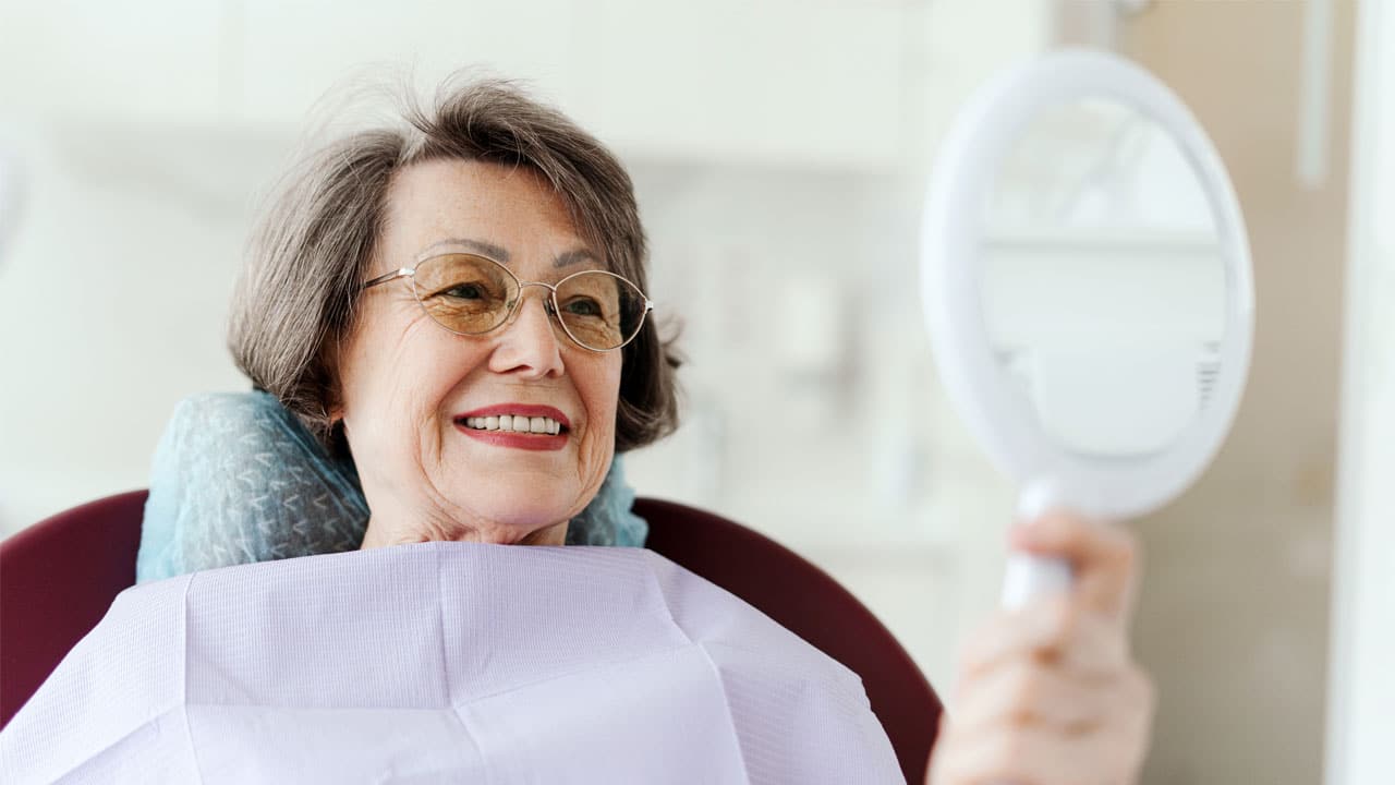 Older dental patient sitting in dental chair looking at teeth in mirror